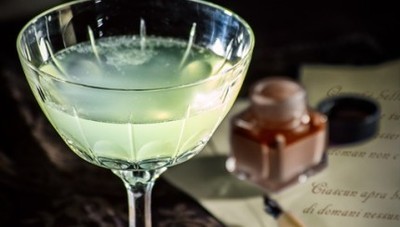 A classic cocktail in a crystal glass.  The glass has a chartreuse-green drink inside, and there is a pen fountain in the background slightly out of focus. 